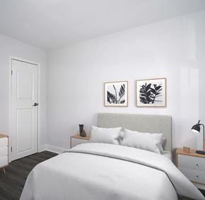Baltimore apartment for rent - A small bedroom with white walls and gray-brown vinyl floors. The bed has a light gray headboard and a white comforter and pillows. Two foliage prints hang on the wall above the headboard. Small end tables are on either side of the bed, and one has a lamp on it. The door is on the wall to the left of the bed.
