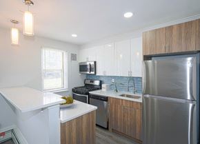Baltimore apartment for rent - kitchen with blue tile backsplash and stainless-steel appliances. The walls are white and the floor is gray-brown vinyl. Cabinets above the fridge are brown and the cabinets under the countertops are brown.