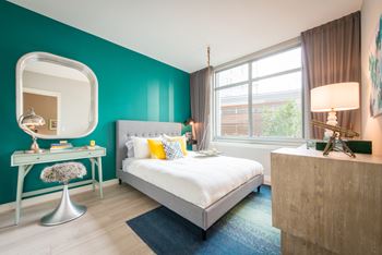 A bedroom with a bed, a desk with a chair, a mirror, and a lamp.