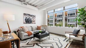 Chicago apartment for rent - A living room in a residence at Wrigleyville Lofts. A couch and coffee table of off to the left, with a small, sectional table to the near left. Out the windows, to the right, other buildings are seen on a bright day.