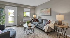 Courtenay apartment for rent - An open living room with a couch, large chair, coffee table and end tables. A patio door with glass insert next to a window and wall mounted AC unit