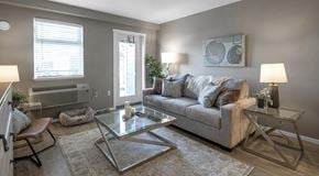 Campbell River apartment for rent - An open living room with a couch, coffee table, chair and end tables. One wall has a balcony door, medium sized window and an AC unit below