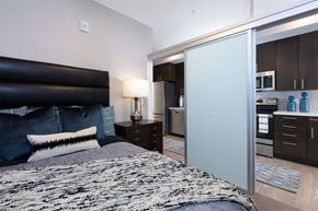 Oakland apartment for rent - a bedroom with a bed and a nightstand with a lamp and a kitchen in the background