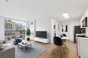 Philadelphia apartment for rent - A modern living room with a grey sofa, a white coffee table, a black chair, a white TV stand, and a kitchen area with a sink and cabinets.