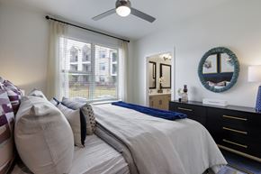 Pflugerville apartment for rent - A bedroom with a large bed, a ceiling fan, and a window with a view of a building outside.
