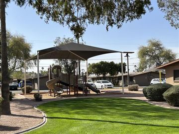 Phoenix apartment for rent - A playground with a slide and a canopy is surrounded by a grassy area and a sidewalk.