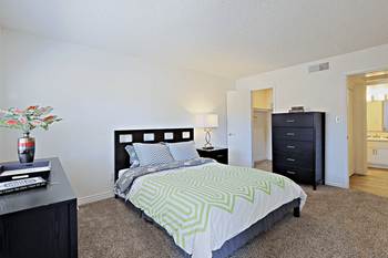 A spacious bedroom with a bed, green patterned bedding, two dressers, and tan carpeting 