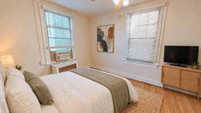 Washington apartment for rent - A bedroom with a bed, a nightstand, a window with blinds, a painting on the wall, and a television on a cabinet.