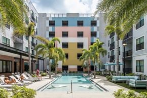 Tampa apartment for rent - A swimming pool is surrounded by palm trees and lounge chairs in front of a colorful building.