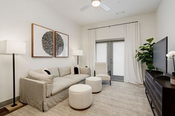 Nashville apartment for rent - A living room with a grey couch, a white ottoman, a painting on the wall, a television, and a plant.