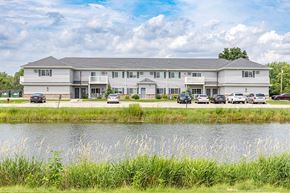 Oshkosh apartment for rent - A large white building with a grey roof is situated by a body of water with greenery in the foreground.