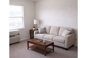 Hopkinsville apartment for rent - A living room with a beige couch, a small table with a lamp, and a window with an air conditioner.