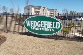 Raeford apartment for rent - A sign for Wedgefield Apartments is shown in front of a fence and some grass.