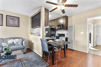 A living room with a table and a kitchen with a ceiling fan