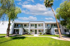 Green Valley apartment for rent - A white building with a balcony on the second floor is surrounded by green grass and trees.