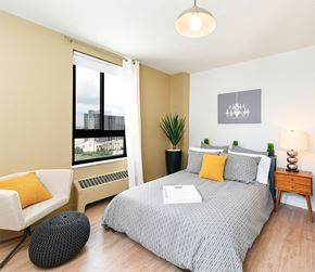 Detroit apartment for rent - A bedroom with a large bed, a chair, and a window with a view of a cityscape.