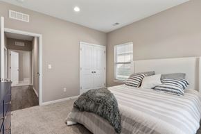 Belton apartment for rent - Senior Housing in Belton, MO - Traditions Villas 55+ - Bedroom with Carpet Flooring, High Ceiling, Light Fixtures, Large Bed, Dresser, Window with Blinds, and Doorway.