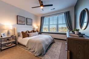 Carmel apartment for rent - North Indianapolis, IN, Apartments Near Palladium - North End Apartments - Primary Bedroom With Plush Bed, Ceiling Fan, Nightstands, Lamps, Dresser, and Wide Window With Curtains.