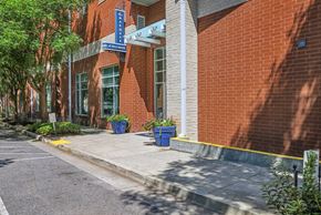 Nashville apartment for rent - 2-Bedroom Apartments in Nashville, TN - The Marquee at Belle Meade - Street View of the Brick Building Entrance With Glass Awning, Property Sign, Blue Planters, And Sidewalk Beside Large Windows.