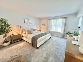 Marlton apartment for rent - A bedroom with a large bed, a nightstand with a lamp, a plant, and a window with curtains.
