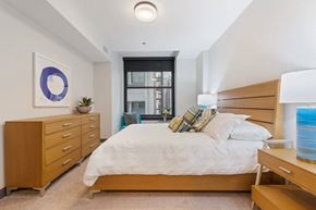 Chicago apartment for rent - A bedroom with a large bed, a wooden dresser, and a window with a view of a cityscape.