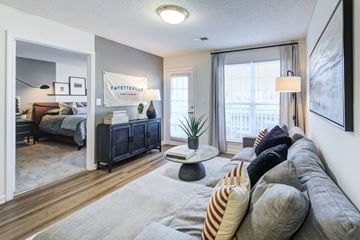 Fayetteville apartment for rent - A living room with a grey sofa and a bedroom with a bed and a nightstand in the background.