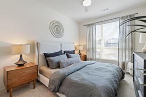 Kansas City apartment for rent - Brookside, Kansas City, MO, Apartments Near Country Club Plaza - Brookside Commons - Bedroom with Carpet Flooring, Large Bed, Nightstands, Lamps, Dresser, Wall Decor, and a Large Window with Blinds and Curtains.