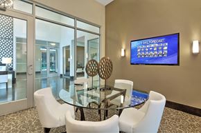 Houston apartment for rent - A modern dining room with a glass table and chairs, a TV displaying a 7 day forecast, and a view of a hallway through the glass doors.