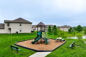 Peculiar apartment for rent - Townhomes in Peculiar, MO - Bradley’s Crossing - An Outdoor Playground Featuring a Play Structure on a Wood Fiber Surface Surrounded by Grass, Trees, Benches, and Townhomes.