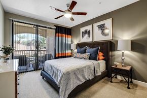 Phoenix apartment for rent - A stylish and inviting bedroom here at Desert Club featuring a spacious bed with layered bedding and accent pillows, flanked by matching nightstands with modern lamps. Large sliding glass doors with vertical blinds and bold drapery panels bring in natural light and provide access to a private patio, while a ceiling fan and soft carpeting add comfort and warmth to the space.