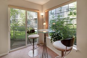 Anchorage apartment for rent - A bright dining nook at Admirals Cove featuring a round glass table with two modern bar-height chairs. Large windows and a sliding glass door overlook green landscaping, filling the space with natural light. Soft carpeting and corner lighting create a relaxed spot for meals or morning coffee.