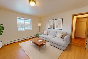 Wasilla apartment for rent - Bright living room at Amber Ridge with wood-style flooring, a large window, and a light gray sectional sofa with neutral pillows. A wooden coffee table sits on a cream area rug, paired with framed abstract art and a floor lamp. An open doorway reveals a walk-in closet, and a potted plant adds greenery.