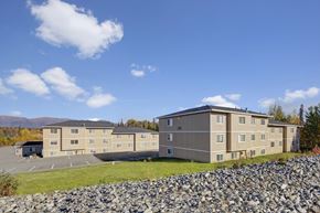 Wasilla apartment for rent - Wide exterior shot of Amber Ridge Apartments showing two beige three-story residential buildings with multiple windows, surrounded by trees with autumn colors and mountains visible in the distance under a bright blue sky.
