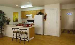Wasilla apartment for rent - Inviting kitchen and entryway at Amber Ridge with wood cabinets, white appliances, and a breakfast bar set with red dishware. Two metal barstools sit at the counter, with décor, wall art, and a potted plant adding warmth. Light wood-look floors flow to a hallway and front door with natural light.