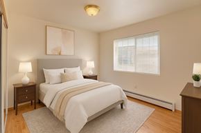 Wasilla apartment for rent - Bright bedroom at Amber Ridge with a neatly made bed featuring white bedding and a beige throw. Two matching nightstands with lamps flank the upholstered headboard, and soft artwork hangs above. A large window fills the room with light, with wood-look floors, a dresser, and a neutral rug completing the space.