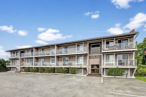 Anchorage apartment for rent - Exterior view of Arbor Pointe apartment building with balconies, large windows, and a parking lot in front under a blue sky with scattered clouds.