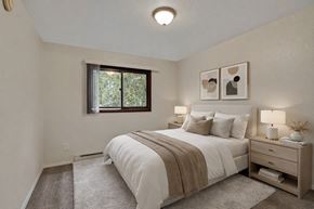 Anchorage apartment for rent - Cozy bedroom at Arbor Pointe with a large bed dressed in white bedding and a tan throw blanket, matching nightstands with lamps, wall art above the bed, and a window bringing in natural light from outside greenery