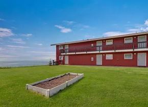 Kenai apartment for rent - A scenic exterior view of Bay Arms Apartments featuring a two-story red building with balconies and white trim. A wide grassy lawn stretches across the property with a raised planting bed in the center. The ocean sits just beyond the grounds, offering peaceful waterfront views.