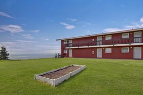 Kenai apartment for rent - A scenic exterior view of Bay Arms Apartments featuring a two-story red building with balconies and white trim. A wide grassy lawn stretches across the property with a raised planting bed in the center. The ocean sits just beyond the grounds, offering peaceful waterfront views.
