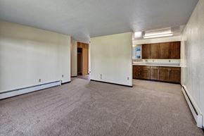 Kenai apartment for rent - A spacious living area at Bay Arms Apartments with plush carpet and neutral walls. The open layout leads into the kitchen, which features wood cabinets and a long countertop. A hallway and closet space sit nearby, offering functional storage and room flexibility.