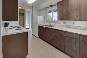 Kenai apartment for rent - A spacious kitchen at Bay Arms Apartments featuring wood cabinets, long countertops, a double sink, and a stovetop. A large refrigerator sits near a bright window with blinds that bring in natural light. Neutral flooring and ample storage make the space functional and convenient.