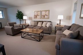 Fairbanks apartment for rent - A spacious living room at Birchwood Apartments featuring matching gray sofas and an armchair with neutral pillows. Two framed artworks hang above the main couch, and end tables with lamps flank the seating. A wooden coffee table and a tall potted plant complete the warm, inviting space.