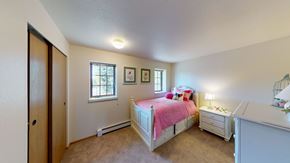 Fairbanks apartment for rent - A cheerful Birchwood bedroom with soft beige walls and carpet, featuring a white twin bed with storage drawers and bright pink bedding topped with playful accent pillows. Two windows let in natural light above floral wall art. A matching nightstand with a lamp and a coordinating dresser complete the cozy space beside a wide closet with sliding wooden doors.