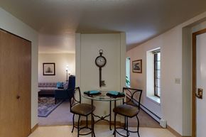 Fairbanks apartment for rent - A cozy dining nook at Birchwood Apartments featuring a round glass table with two cushioned chairs set against a dividing wall decorated with a vintage key-shaped wall clock, with views into the adjoining living room and hallway lined with windows.