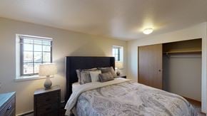 Fairbanks apartment for rent - A bright Birchwood bedroom featuring a dark upholstered headboard with paisley bedding and layered pillows centered against a neutral wall. Two matching lamps on wooden nightstands sit beside the bed beneath twin windows. The room includes soft carpet and a wide closet with sliding wooden doors partially open.