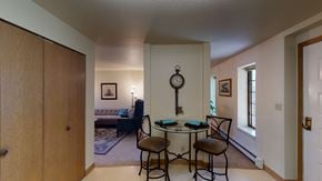 Fairbanks apartment for rent - A cozy dining nook at Birchwood Apartments featuring a round glass table with two cushioned chairs set against a dividing wall decorated with a vintage key-shaped wall clock, with views into the adjoining living room and hallway lined with windows.