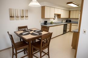 Fairbanks apartment for rent - Dining space at Birchwood Apartments featuring a wooden table set for four with rolled napkins, opening to a spacious kitchen with light cabinetry, long countertops, and modern appliances.