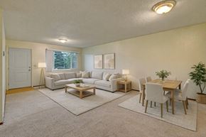 Anchorage apartment for rent - A spacious Brighton Woods living and dining area with soft neutral décor, featuring a large light gray sectional sofa, two coordinating area rugs, and a wooden coffee table. A matching dining set for six sits nearby, accented by a potted plant and simple wall art, all illuminated by warm overhead lighting and a bright window view.