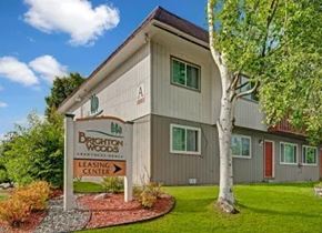 Anchorage apartment for rent - Lush Brighton Woods apartment community exterior on a sunny day featuring a two-story beige and green building framed by mature trees and manicured landscaping. A prominent leasing center sign with orange lettering stands near the front walkway, bordered by neat red rock mulch, shrubs, and a bright green well-kept lawn.