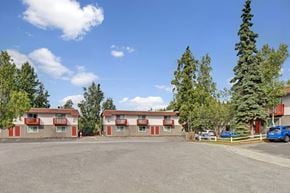 Anchorage apartment for rent - A sunny Brighton Woods community courtyard with two matching two-story apartment buildings featuring tan siding, red trim doors, and small balconies facing a wide parking area. A few cars are parked to the right, and tall evergreens and leafy trees frame the property beneath a bright blue sky dotted with soft white clouds.